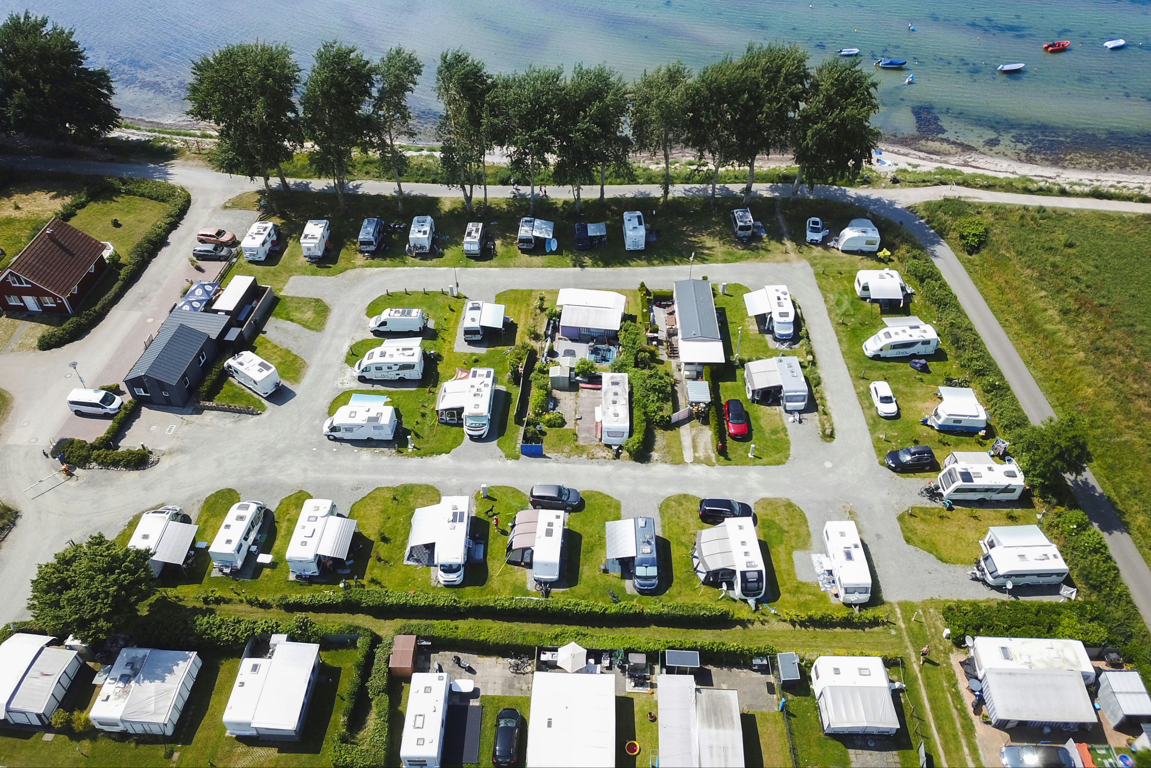 Luftaufnahme eines Campingplatzes mit vielen Wohnmobilen und Stellplätzen, Wegen, Rasenflächen und Bäumen am Ufer eines Sees.