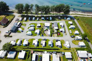 Luftaufnahme eines Campingplatzes mit vielen Wohnmobilen und Stellplätzen, Wegen, Rasenflächen und Bäumen am Ufer eines Sees.