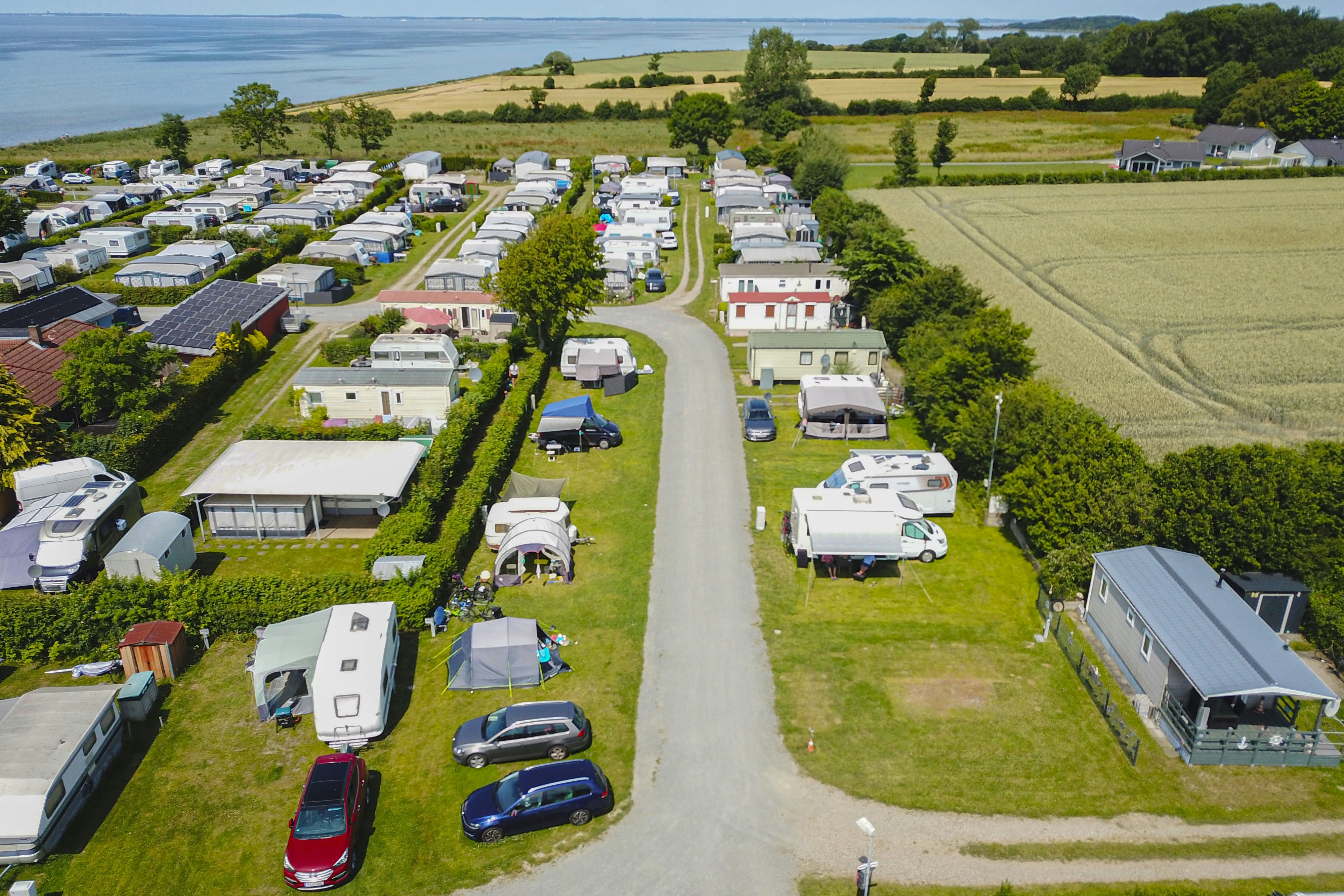 Luftaufnahme eines Campingplatzes mit Wohnmobilen und Stellplätzen entlang eines breiten Weges, umgeben von Feldern und Bäumen am See.