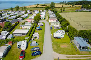 Luftaufnahme eines Campingplatzes mit Wohnmobilen und Stellplätzen entlang eines breiten Weges, umgeben von Feldern und Bäumen am See.