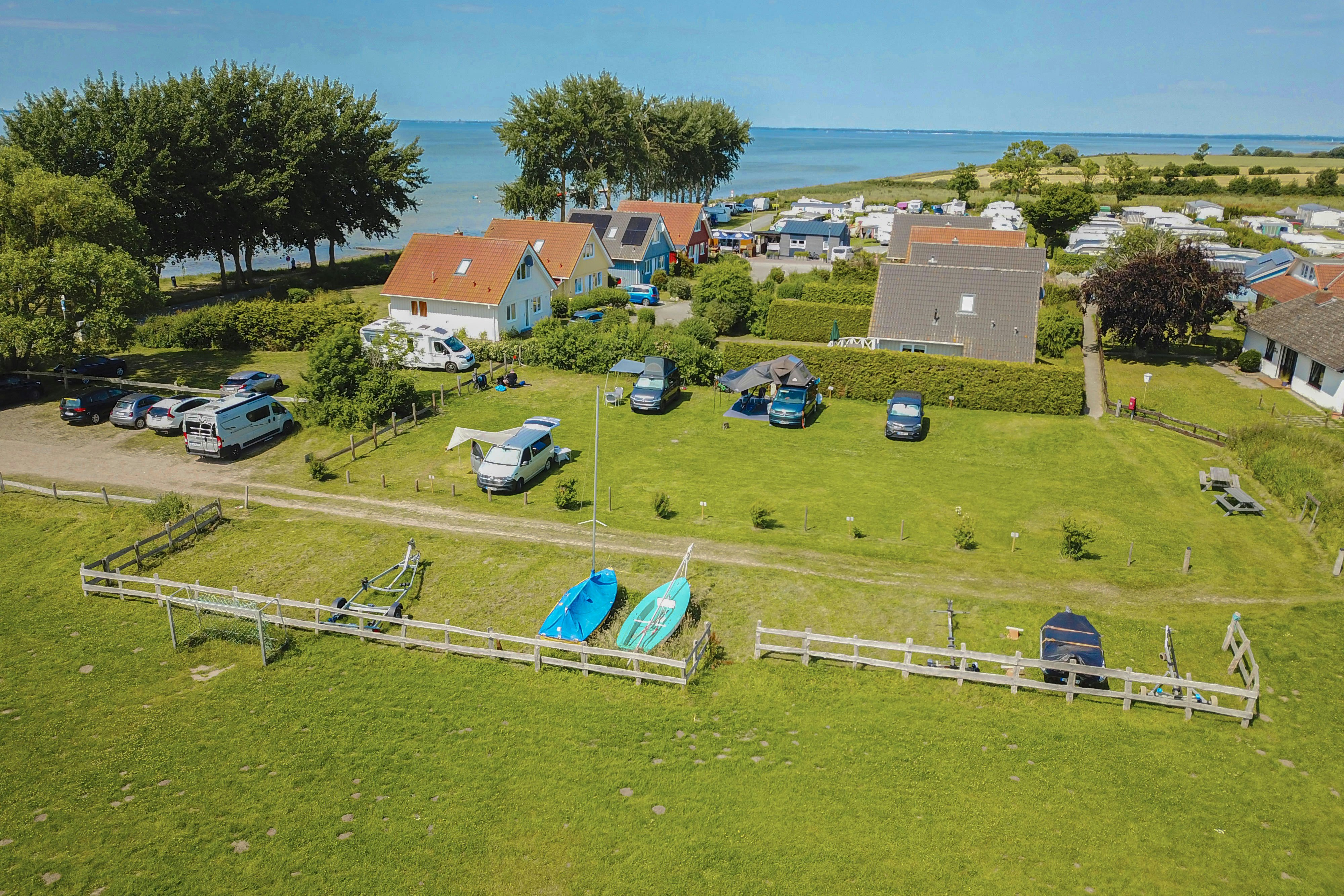 Luftaufnahme von Campinghäusern und Wohnwagen auf einer grünen Anlage, mit Rasenflächen, Wegen, Hecken und Blick auf das Meer.