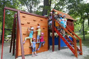 Kinder spielen am hölzernen Spielhaus mit Rutsche auf dem Campingplatz, im Hintergrund Bäume und ein Kiesweg.