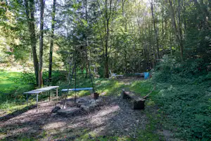 Campingplatz im Wald: Feuerstelle mit Grillrost, Holzbänke und Tisch, umgeben von Bäumen und grünem Unterholz.