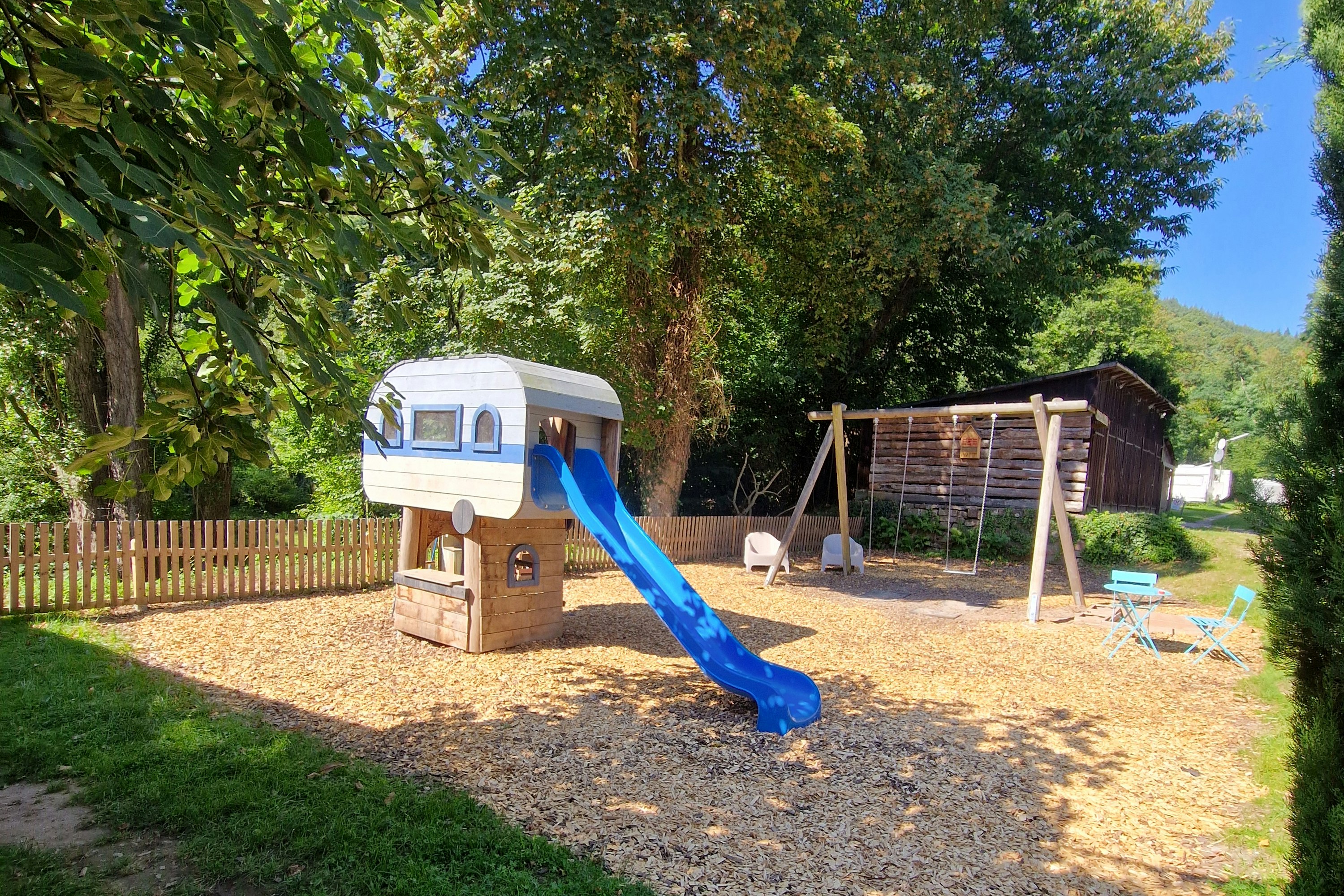 Wohnwagen mit blauem Rutschbahn-Spielbereich auf dem Campingplatz, umgeben von Bäumen und Rasen, mit Schaukelgestell und Sandfläche.