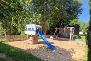 Wohnwagen mit blauem Rutschbahn-Spielbereich auf dem Campingplatz, umgeben von Bäumen und Rasen, mit Schaukelgestell und Sandfläche.