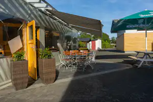 Terrasse am Zelt mit Holz-Tisch und Stühlen, Sichtschutz und gelber Tür, daneben Holzhaus und grüner Sonnenschirm.