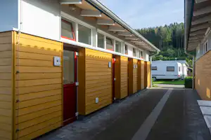 Reihenhausartige Camping-Unterkunft mit Holzfassade, roten Türen und Fenstern, gepflasterter Zufahrt und Wohnwagen im Hintergrund.