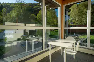 Terrasse mit weißem Tisch und Stühlen vor großen Fenstern, Blick auf Wohnwagen und Bäume im Campingbereich.