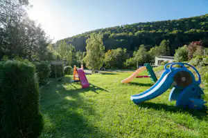 Spielplatz mit blauer Rutsche und Kletterelementen auf einer grünen Wiese, im Hintergrund Gebäude und Waldhang.