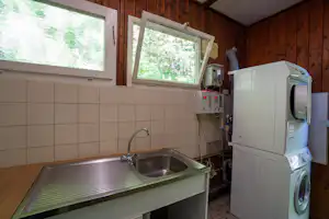 Waschraum mit Spülbecken, Armatur und Waschmaschine, dahinter Fliesenwand und Fenster mit Blick ins Grüne.