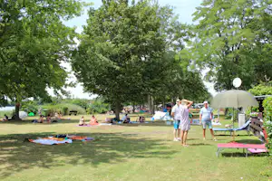 Campingwiese mit mehreren Zelten, Liegestühlen und Personen zwischen Bäumen, im Hintergrund ein weißes Zelt und Spieldecken.