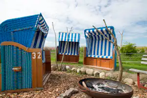 Blaue Strandkörbe und ein Grill auf einem Campingplatz, mit Holzverkleidung, Kiesboden und Steinen im Vordergrund.