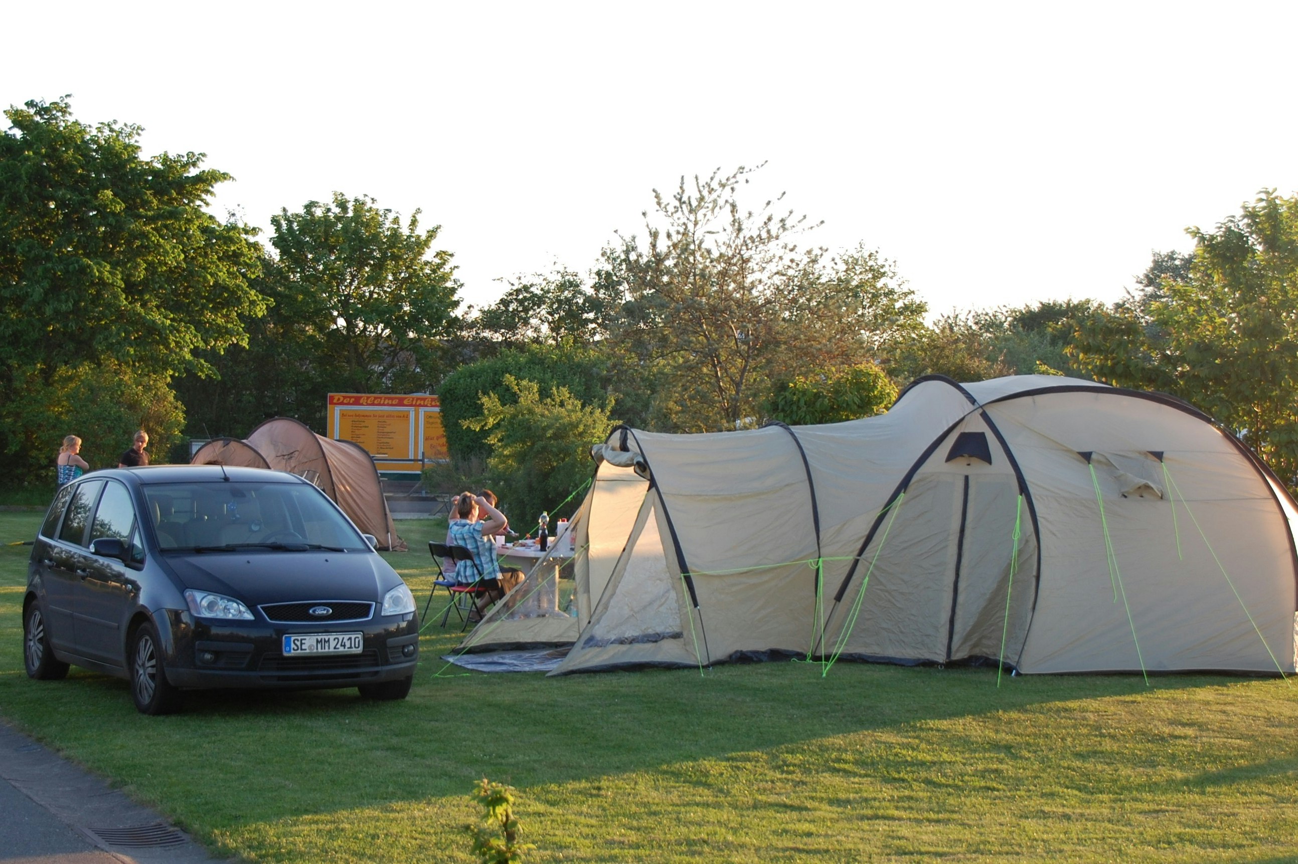 Campingzelt auf einer Wiese, daneben ein Auto und ein kleiner Tisch mit Stühlen, im Hintergrund Bäume und ein Weg.