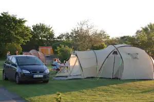 Campingzelt auf einer Wiese, daneben ein Auto und ein kleiner Tisch mit Stühlen, im Hintergrund Bäume und ein Weg.