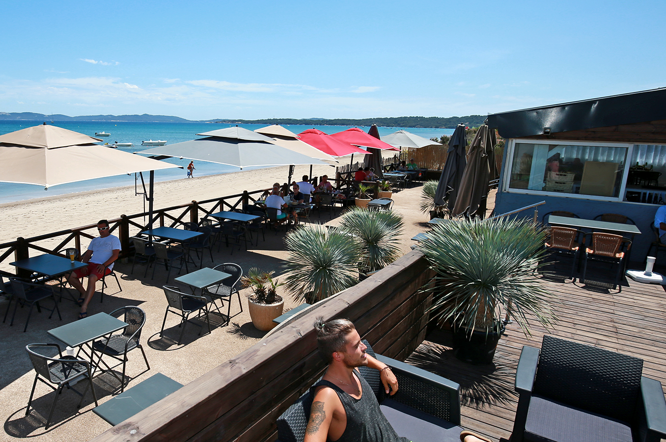 Terrasse am Campingplatz am Meer mit Sonnenschirmen, Holzzaun, Sitzgruppen und Blick auf den Strand und das Wasser.