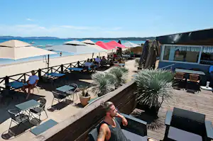 Terrasse am Campingplatz am Meer mit Sonnenschirmen, Holzzaun, Sitzgruppen und Blick auf den Strand und das Wasser.