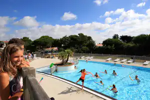 Familie am Camping-Pool: Kinder und Erwachsene im Wasser, Liegen am Beckenrand, Bäume und Gebäude im Hintergrund.