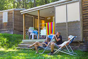 Mobilheim mit Holzfassade und Terrasse im Grünen, zwei Liegestühle vor dem Eingang, bunte Decke am Geländer.