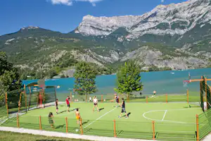 Sportplatz am Campingplatz am See, Basketballkorb und Spieler auf dem Kunstrasen, im Hintergrund Berge und türkisfarbenes Wasser.