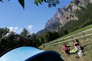 Campingzelt am Bergpfad, daneben sitzen zwei Personen mit Campingstühlen und Tisch vor grünen Hängen und Felsbergen.