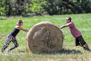 Zwei Personen rollen einen Strohballen auf einer grünen Wiese im Campingbereich, im Hintergrund Bäume.