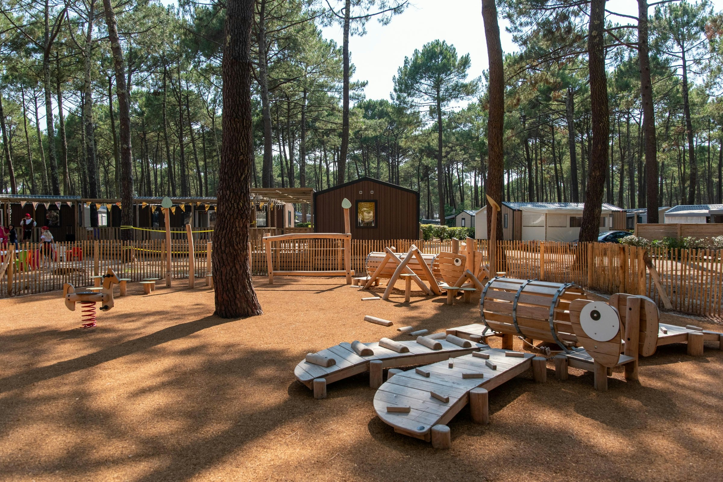 Holzterrasse mit Sitz- und Liegestühlen, daneben ein Fasssauna-ähnliches Holzfass, umgeben von Kiefern im Campingbereich.