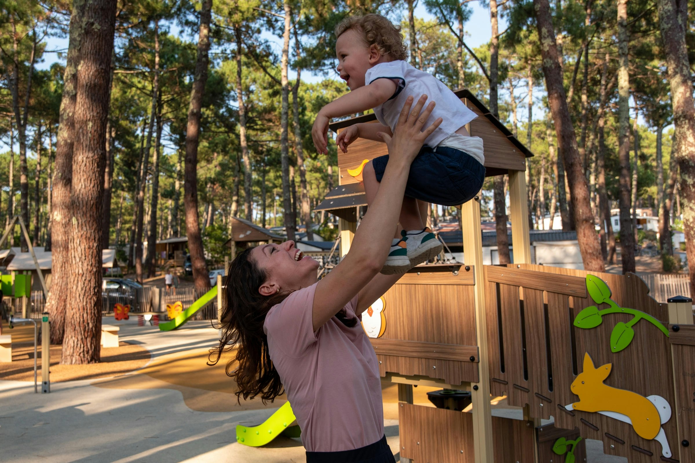 Ein Kind hebt ein anderes Kind an einem Spielbereich im Campingplatz zwischen hohen Bäumen, mit Holzelementen und bunten Spielzeugteilen.