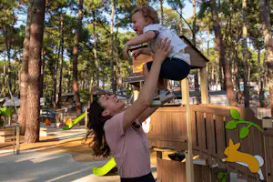 Ein Kind hebt ein anderes Kind an einem Spielbereich im Campingplatz zwischen hohen Bäumen, mit Holzelementen und bunten Spielzeugteilen.