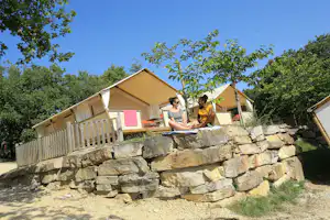 Glamping-Zelt mit Terrasse, zwei Personen sitzen auf einer Decke vor dem Zelt, umgeben von Bäumen und Steinmauer.