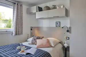 Bett in einem modernen Camping-Unterkunftsraum mit Kissen, Nachttisch, Regalen und Vorhang am Fenster.