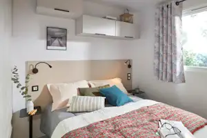 Bett mit Kissen und Decke in einem hellen Wohnbereich, daneben Nachttisch und Fenster mit gemusterten Vorhängen.