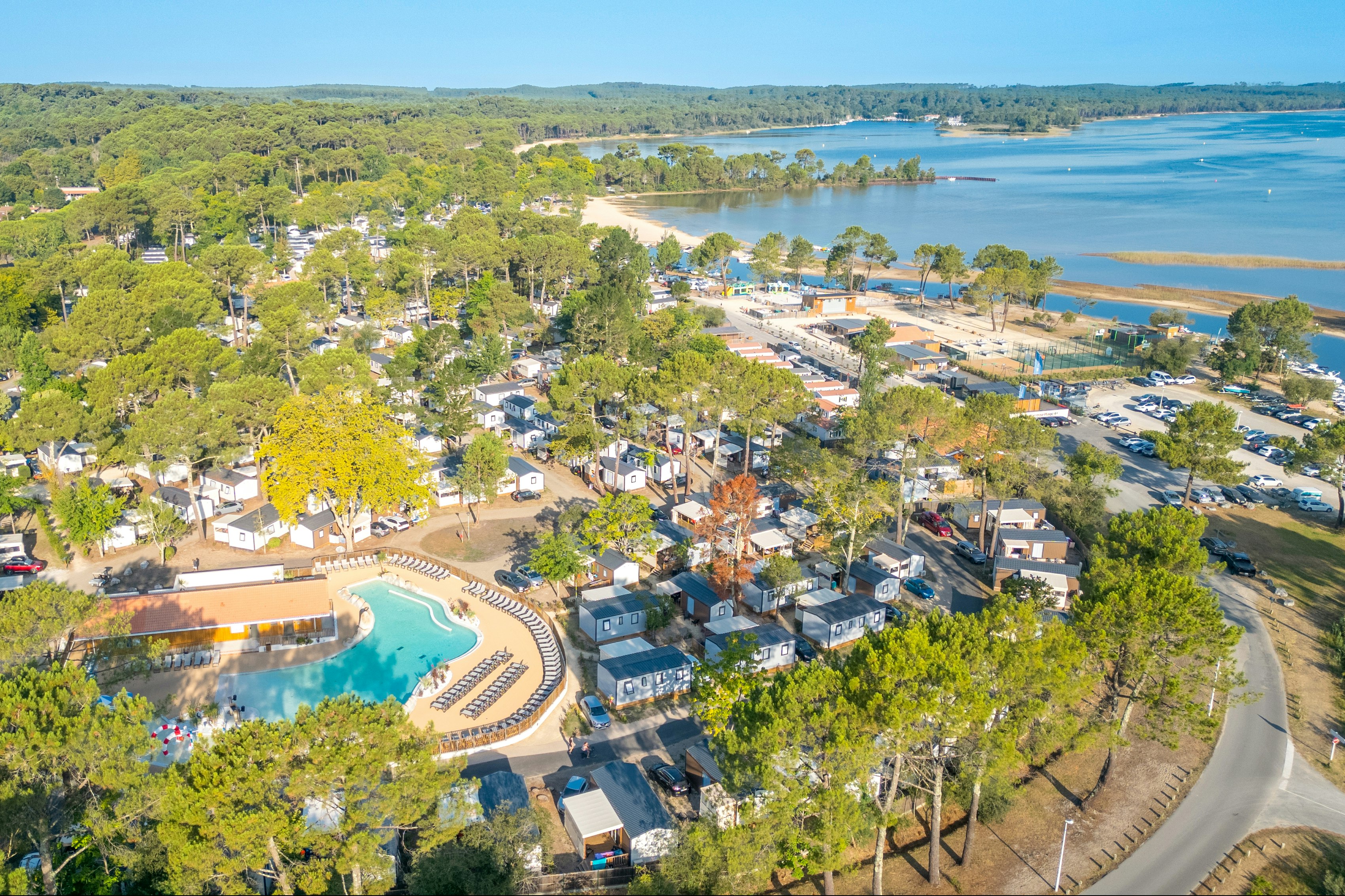 Luftaufnahme eines Campingplatzes mit Stellplätzen, kleinen Häusern und einem Poolbereich am Fluss, umgeben von Bäumen und Wegen.