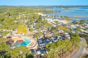 Luftaufnahme eines Campingplatzes mit Stellplätzen, kleinen Häusern und einem Poolbereich am Fluss, umgeben von Bäumen und Wegen.