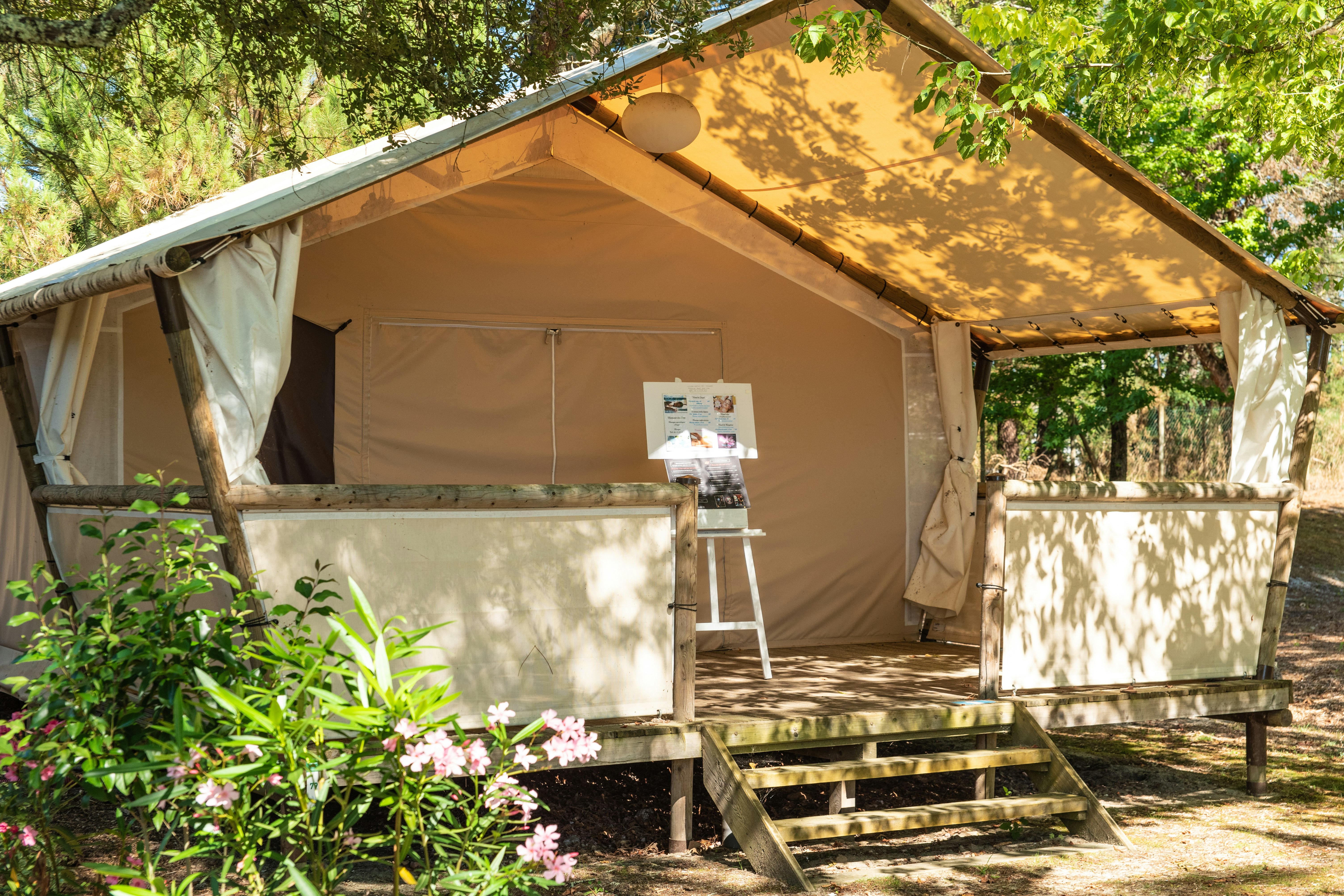Zelt mit Holzpodest und Stufen, Sonnensegel über dem Eingang, umgeben von Büschen und Blumen im Campingbereich.