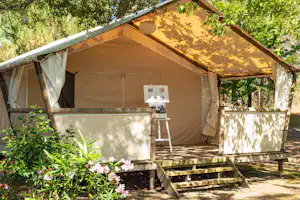 Zelt mit Holzpodest und Stufen, Sonnensegel über dem Eingang, umgeben von Büschen und Blumen im Campingbereich.