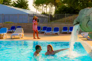 Kinder am Pool mit Wasserspiel, blaue Liegen und Sonnenschirme, im Hintergrund Zeltplane und Bäume.