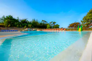 Campingpool mit türkisfarbenem Wasser, Liegen am Beckenrand, Zaun und grünen Bäumen im Hintergrund.