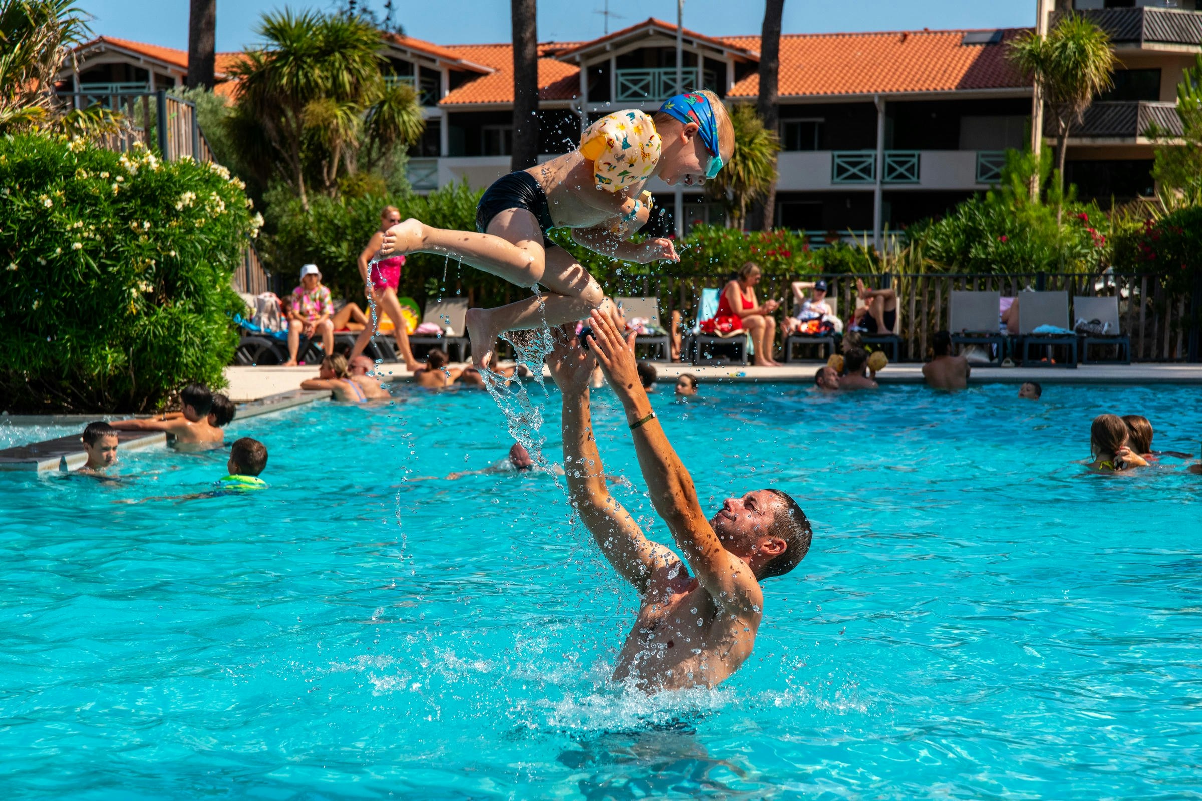 Kinder planschen im blauen Pool eines Campingplatzes, im Hintergrund Palmen und Gebäude mit roten Ziegeldächern.