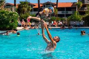 Kinder planschen im blauen Pool eines Campingplatzes, im Hintergrund Palmen und Gebäude mit roten Ziegeldächern.