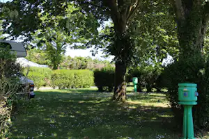 Campingplatz mit grünem Rasen, Bäumen und Hecken; links ein Wohnwagen, rechts ein grüner Abfalleimer am Weg.