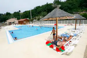 Camping-Pool mit Sonnenschirmen, Liegen und mehreren Personen im Wasser, im Hintergrund Gebäude und Bäume.