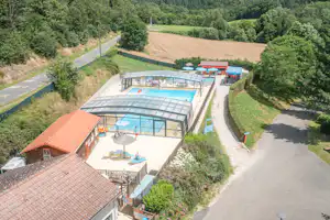 Luftaufnahme eines Campingplatzes mit überdachtem Schwimmbecken, Gebäuden und Wegen zwischen grünen Bäumen und Feldern.