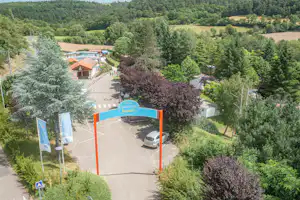 Blick auf den Camping-Eingang mit Torbogen, breitem Weg und gepflegten Bäumen in grüner Landschaft.