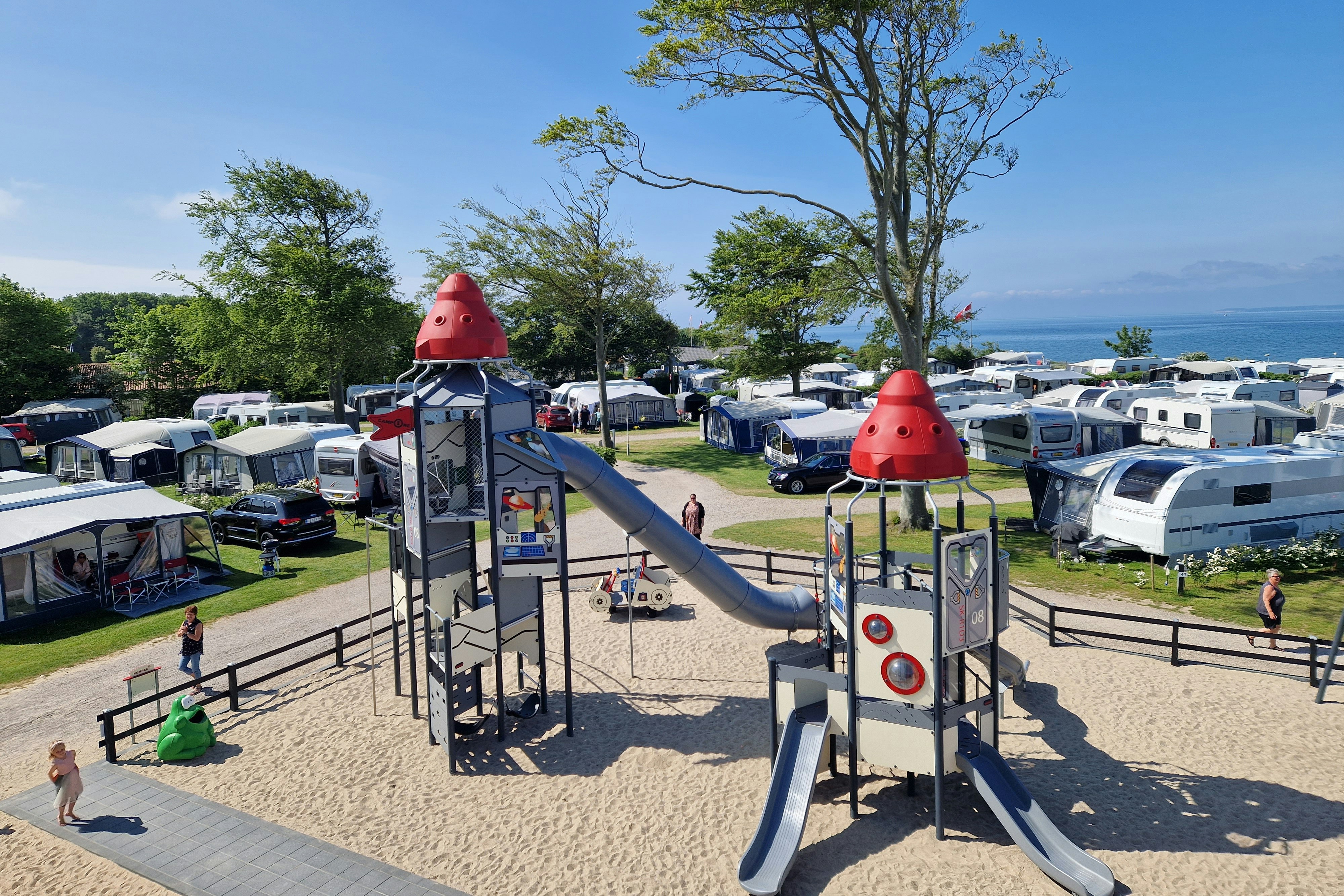 Spielplatz mit Rutsche und Kletterelementen auf dem Campingplatz, im Hintergrund Wohnwagen, Bäume und ein Blick auf das Wasser.