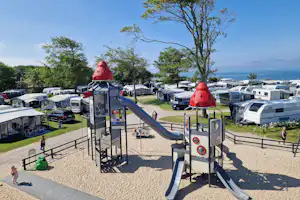 Spielplatz mit Rutsche und Kletterelementen auf dem Campingplatz, im Hintergrund Wohnwagen, Bäume und ein Blick auf das Wasser.