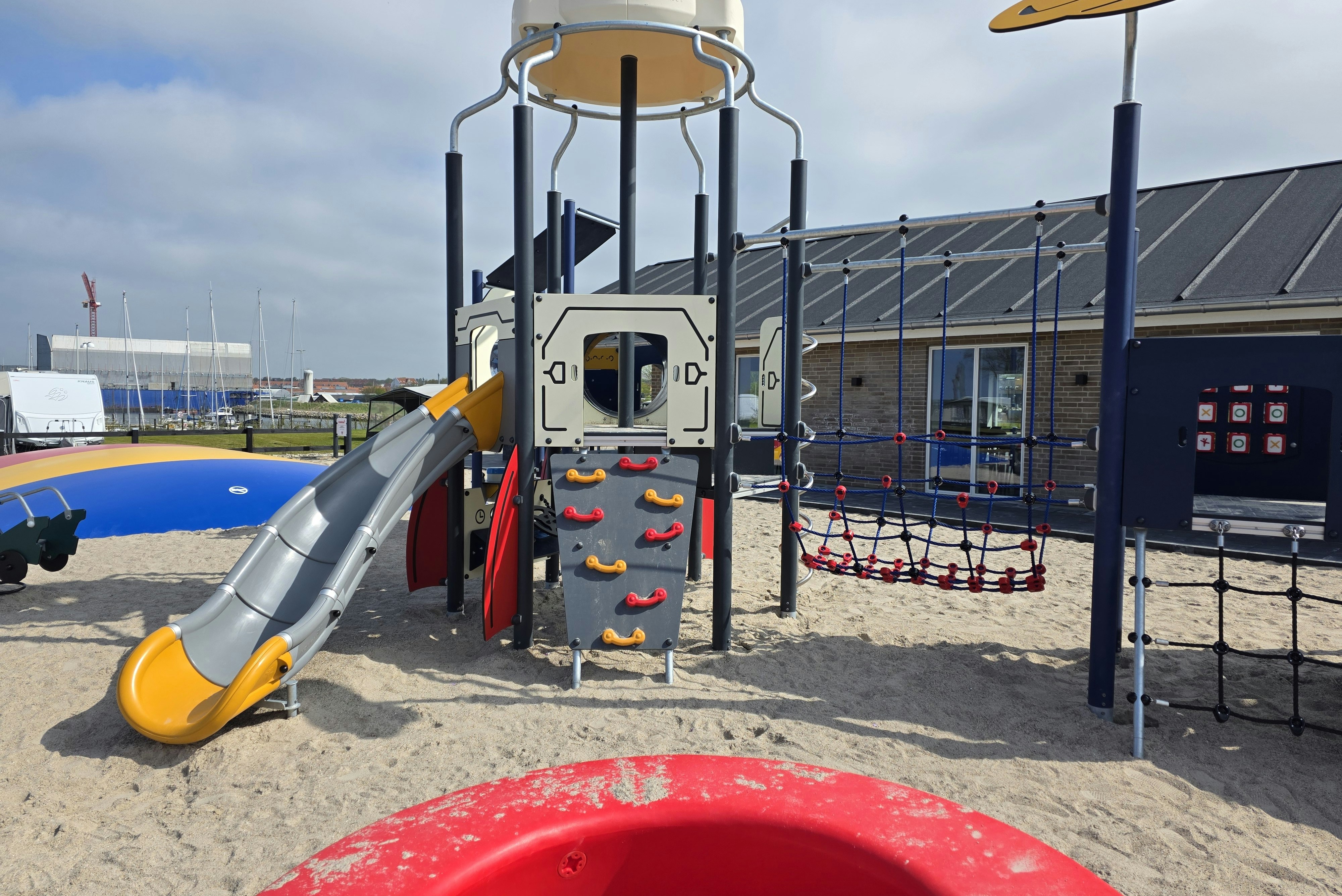 Spielplatz mit Klettergerüst, Rutsche und Netz am Sandboden, im Hintergrund Gebäude und Wohnwagenstellplatz.