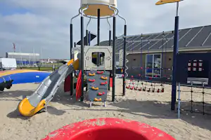 Spielplatz mit Klettergerüst, Rutsche und Netz am Sandboden, im Hintergrund Gebäude und Wohnwagenstellplatz.