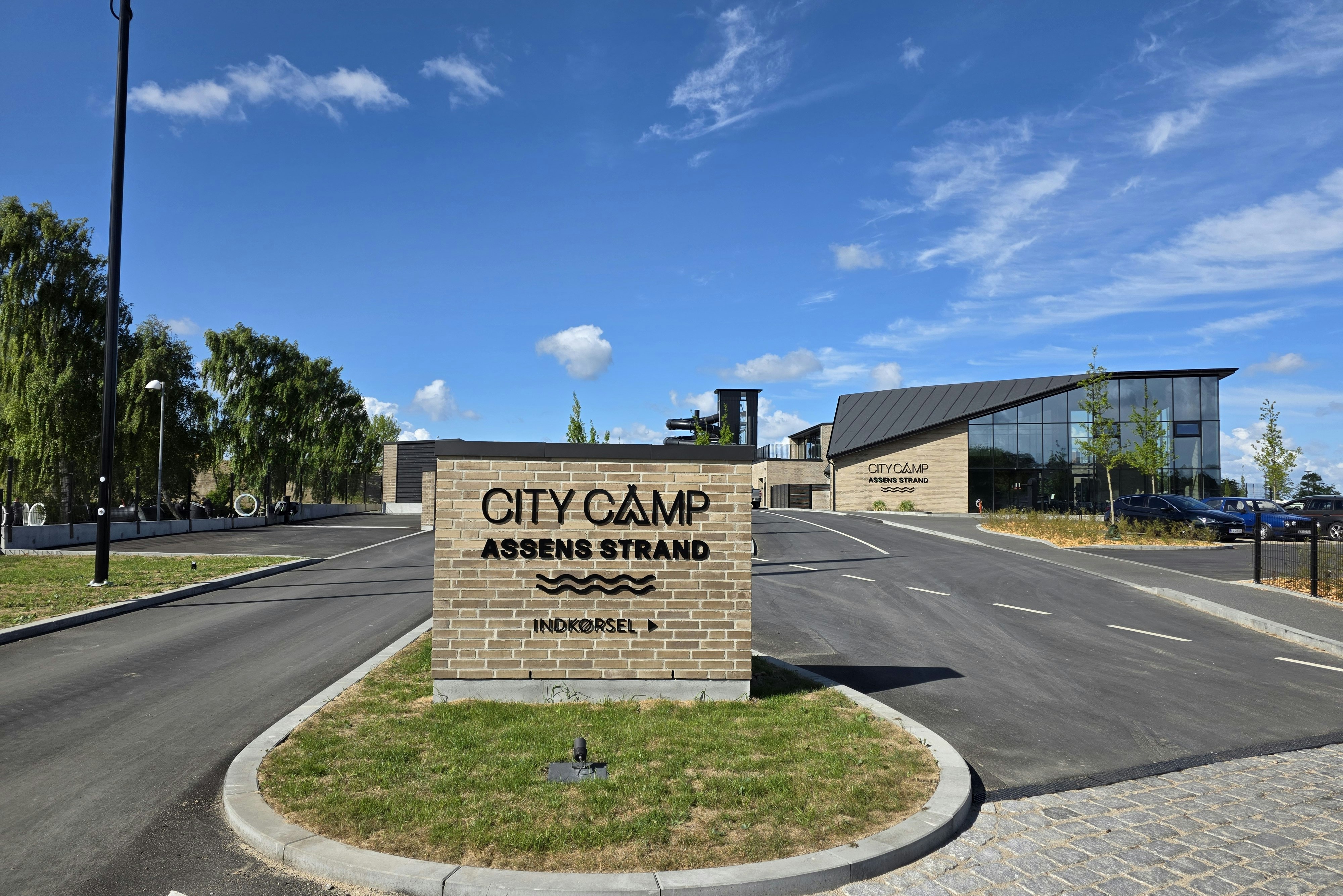 Schild „CITY CAMP“ am Eingang „ASSENS STRAND“ neben Straße und Gehweg, mit Gebäuden und Bäumen im Hintergrund.