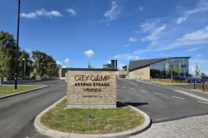 Schild „CITY CAMP“ am Eingang „ASSENS STRAND“ neben Straße und Gehweg, mit Gebäuden und Bäumen im Hintergrund.