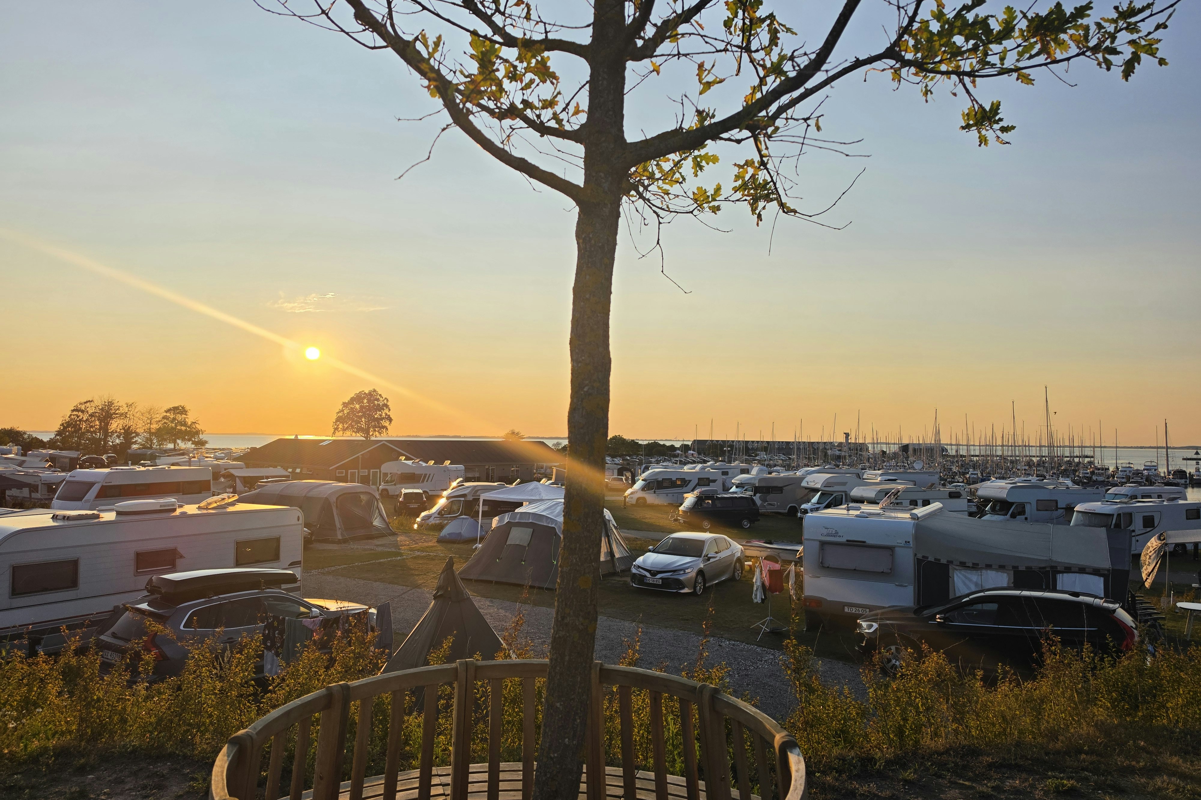 Blick auf einen Campingplatz mit Wohnwagen und Stellplätzen, im Vordergrund eine Holzbank, im Hintergrund ein See bei Sonnenuntergang.